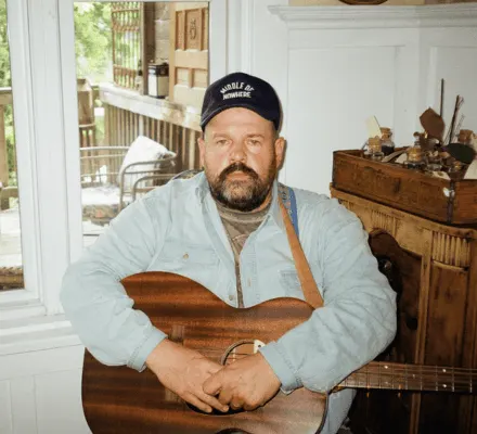 Donovan Woods wears a baseball cap and jean jacket. He holds an acoustic guitar in his lap.