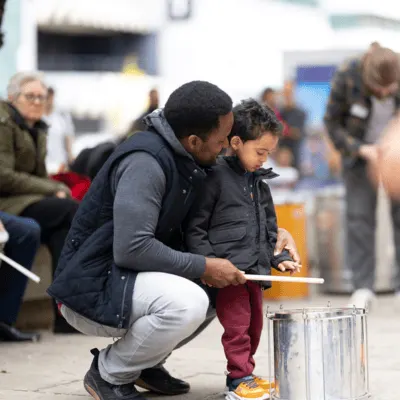 In a busy room with people and instruments, a father squats behind his young son, showing him how to play a snare drum.
