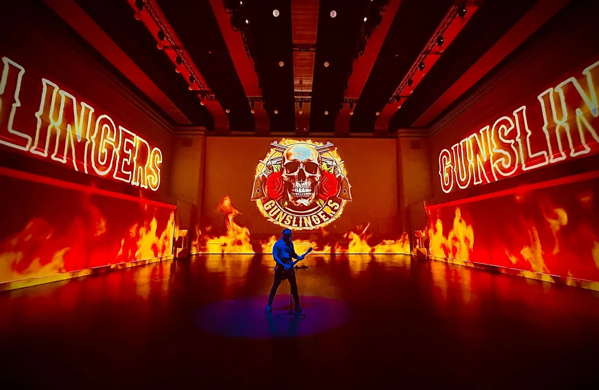 A guitarist plays against bright floor to ceiling screens, with flames on the background.