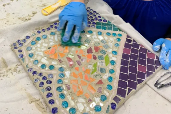 Gloved hand wipes grout into a mosaic panel piece with a heart at the centre of the design.